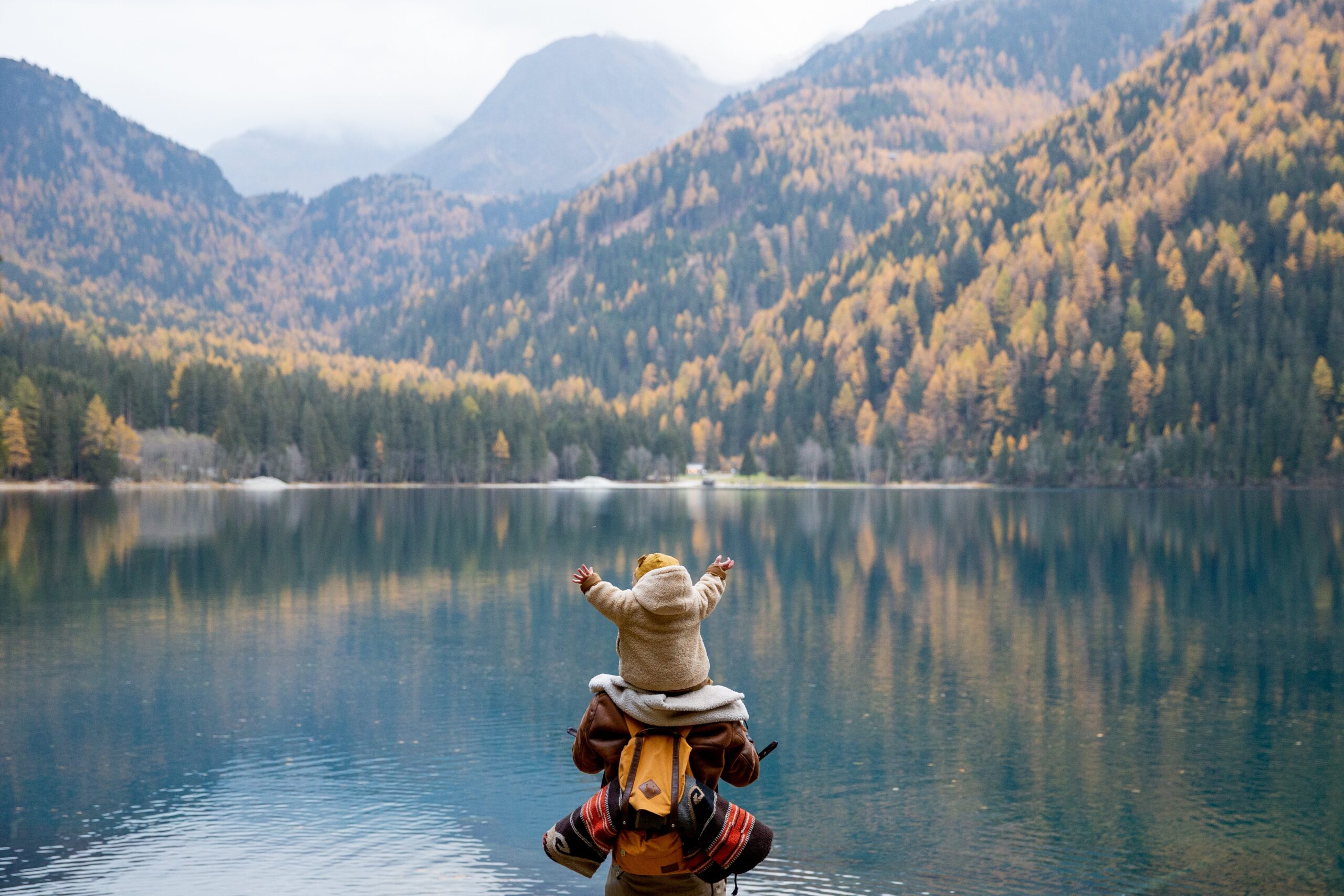 Baby on dad's shoulders. Back to the camera. They are traveling and they are facing a beautiful lake and some yellow flowery mountains